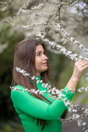 Woman wearing a green top and white pants. She stands beside a bush adorned with delicate white flowers, embracing the essence of spring.