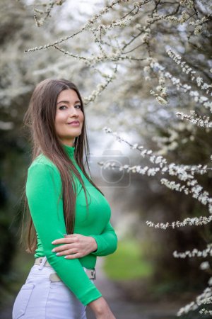 Woman wearing a green top and white pants. She stands beside a bush adorned with delicate white flowers, embracing the essence of spring.