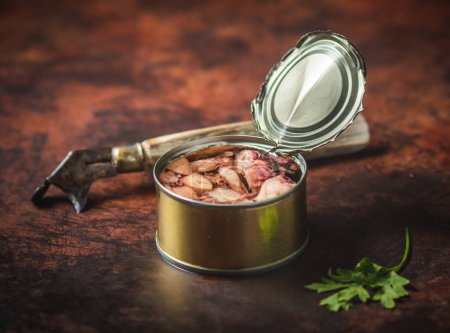 Photo for Canned octopus open on a dark brown table with an old can opener and parsley. Ready for eat. - Royalty Free Image