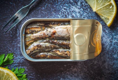 Canned sardines with olive oil open on a dark blue table with a fork, lemon and parsley. Ready for eat. 