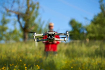 Téléchargez les photos : Nurtingen, Allemagne - 29 mai 2021 : Drone dji air 2s survole un champ de fleurs sauvages. Pilote flou avec veste rouge contrôle le système d'aéronef sans pilote. Arbres verts en arrière-plan. Vue de face. - en image libre de droit