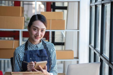 Eine asiatische Geschäftsfrau genießt sein Online-KMU-Geschäft, das Produkte verkauft, von der Eröffnung eines Online-Shops profitiert und Bestellungen für Kundenpakete im Büro entgegennimmt. Konzept KMU.