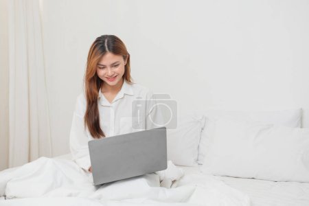 Woman in white nightgown waking up on weekend morning resting and relaxing playing with laptop mobile phone Eating bread and drinking tea in glass inside white bedroom.  Morning vacation concept. Poster 659027570
