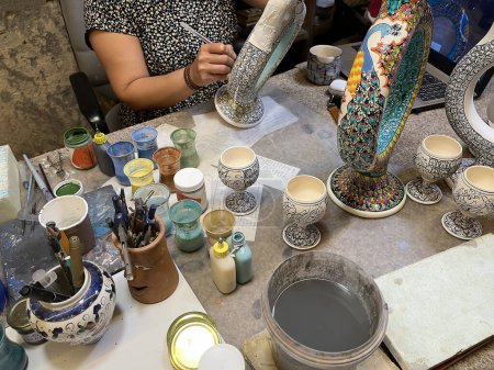 Nahaufnahme eines Arbeitstisches mit Farben und Glasuren zur Verzierung hethitischer Krüge und typisch türkischer Keramiktassen in einer Bastelwerkstatt, eine Kunsthandwerkerin malt dekorative Details auf einen Krug, waagerecht