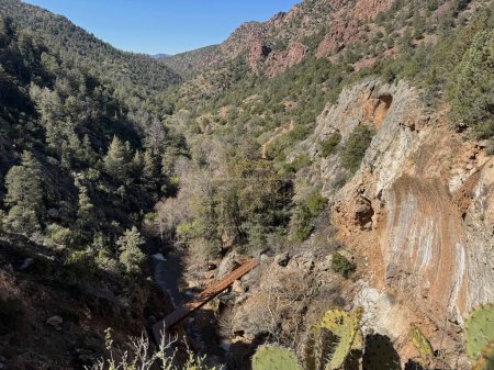 Springtime view of the red rocky mountain landscape and valley in Tonto Natural Bridge State Park in Pine, Arizona with bright blue sky copy space highlighting the lower walking bridge over pine creek leading to the natural bridge. 
