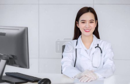 Photo for Smiling Asian professional female doctor sitting in examination room at hospital while on table with paper and computer in health concept - Royalty Free Image