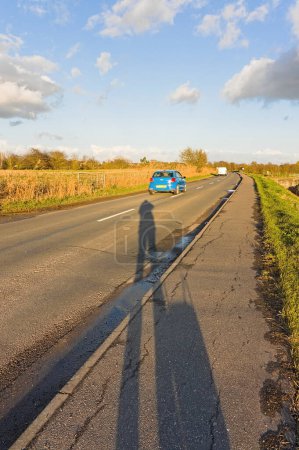 Téléchargez les photos : Ombre d'une personne sur une route de campagne avec une voiture au loin - en image libre de droit