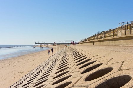 A concrete sea barrier & promenade with people along the beach with a blue sky. Skegness UK