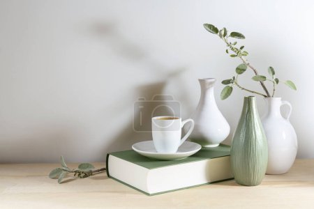 Téléchargez les photos : Still live of three vases with a sage leaf branch, gray green book and a coffee cup on a wooden desk or table against a light gray wall, tranquil home decoration, copy space, selected focus - en image libre de droit
