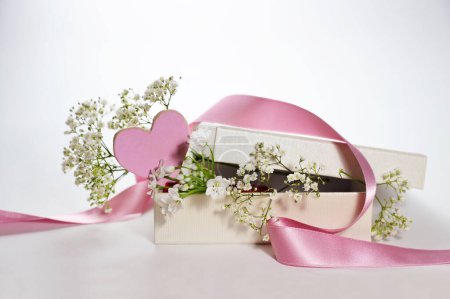 Gift box with a pink painted heart shape, a ribbon and white gypsophila flowers as romantic greeting card for holidays, light background, copy space, selected focus, narrow depth of field