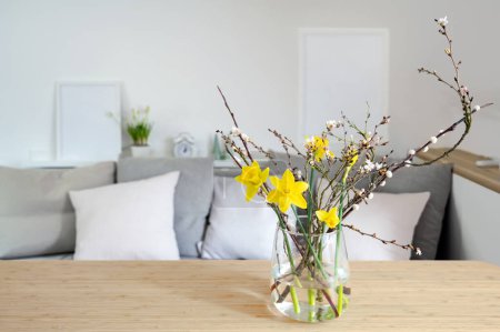 Photo for Spring bouquet with daffodils and flowering branches in a glass vase on a table in front of a sofa bench, seasonal home decor, copy space, selected focus, narrow depth of field - Royalty Free Image