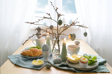 Breakfast table with hanging Easter eggs on spring branches, candles, and blue gray decoration in front of a window with white curtains, copy space, selected focus, narrow depth of field