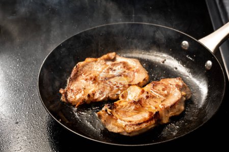 Photo for Pork steak is fried in hot splattering and steaming oil in a black iron skillet on the stove top for a rustic meat dish, copy space, selected focus, narrow depth of field - Royalty Free Image