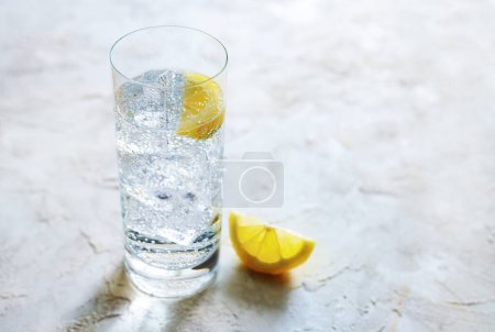 Agua mineral carbonatada fresca con cubitos de hielo y rodajas de limón en un vaso de agua potable, bebida refrescante sobre un fondo de piedra clara, espacio para copiar, enfoque seleccionado, estrecha profundidad de campo 