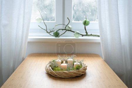 Einfacher Adventskranz mit Moos und weißen Kerzen, die erste wird entzündet, auf einem Tisch am Fenster an einem verschneiten Wintertag, Ferienhausdekoration, Kopierraum, ausgewählter Fokus, enge Schärfentiefe 