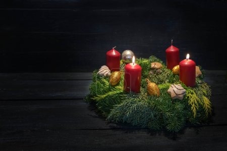 Licht im Dunkeln am zweiten Advent, naturgrüner Kranz mit roten Kerzen, zwei brennen, Weihnachtsdekoration und Plätzchen, dunkler Holzgrund, Kopierraum, ausgewählter Schwerpunkt