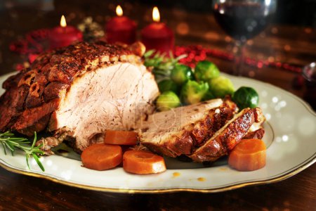 Weihnachtsschinken oder Schweinebraten mit knuspriger Fettkruste, serviert mit Gemüse auf einem dunklen Holztisch mit Kerzen und Bokeh-Blasen, ausgewählter Fokus, enge Schärfentiefe