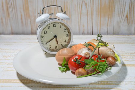 Interval fasting as a healthy diet concept for losing weight, white plate filled to one third with food and an alarm clock on a light rustic wooden background, copy space, selected focus
