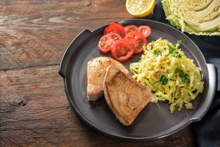 Fried tuna steaks with savoy cabbage and tomatoes on a dark plate and a rustic wooden table, copy space, selected focus, narrow depth of field 