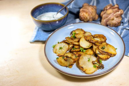 Crispy fried slices of Jerusalem artichoke or topinambur (Helianthus tuberosus) with onions and parsley on a blue plate with a fresh dip, healthy root vegetable, copy space, selected focus