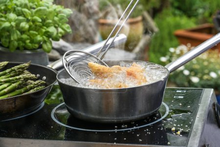 Slotted spoon takes golden deep-fried chicken pieces from a steaming pan with bubbling boiling oil, outdoor kitchen stove surrounded by potted herbs and vegetables, copy space, selected focus 