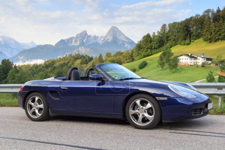 Foto de Berchtesgaden, Alemania - 25 de julio de 2021: Porche Porsche Boxster 986 azul con vista panorámica de la montaña Watzmann y la niebla. El coche es un coche deportivo de dos plazas de motor medio fabricado por Porsche. - Imagen libre de derechos