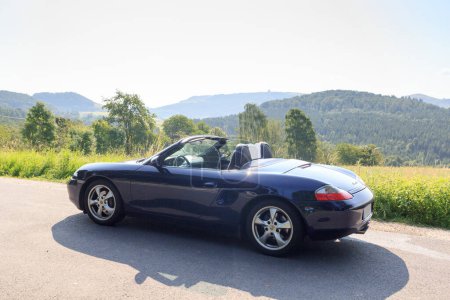 Foto de Gersfeld, Germany - July 23, 2021: Blue roadster Porsche Boxster 986 with Wasserkuppe panorama in Rhoen Mountains. The car is a mid-engine two-seater sports car manufactured by Porsche. - Imagen libre de derechos