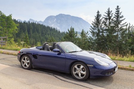 Foto de Berchtesgaden, Germany - July 24, 2021: Blue roadster Porsche Boxster 986 with mountain Hoher Goell panorama at Rossfeldpanoramastrasse (horse field panoramic motorway). It is a sports car by Porsche. - Imagen libre de derechos