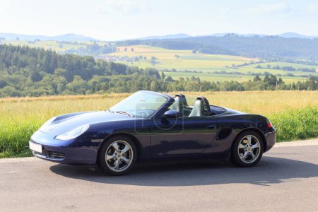 Photo for Gersfeld, Germany - July 23, 2021: Blue roadster Porsche Boxster 986 with Rhoen Mountains panorama. The car is a mid-engine two-seater sports car manufactured by Porsche. - Royalty Free Image