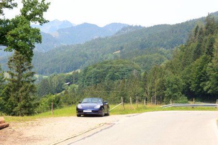 Foto de Bayrischzell, Germany - July 24, 2021: Blue roadster Porsche Boxster 986 with Bavarian Alps panorama at German Alpine Road. The car is a mid-engine two-seater sports car manufactured by Porsche. - Imagen libre de derechos
