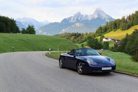 Photo for Berchtesgaden, Germany - July 25, 2021: Blue roadster Porsche Boxster 986 with mountain Watzmann and fog panorama. The car is a mid-engine two-seater sports car manufactured by Porsche. - Royalty Free Image