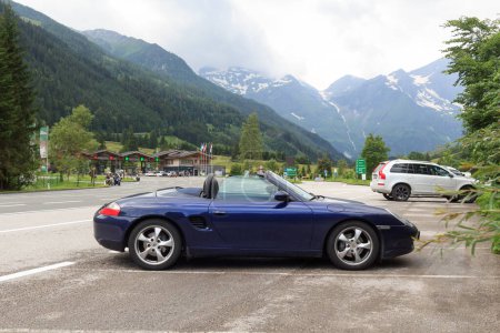 Foto de Fusch, Austria - 25 de julio de 2021: El roadster azul Porsche Boxster 986 con panorama de montaña y la cabina de peaje Grossglockner High Alpine Road. El coche es un coche deportivo de dos plazas fabricado por Porsche. - Imagen libre de derechos