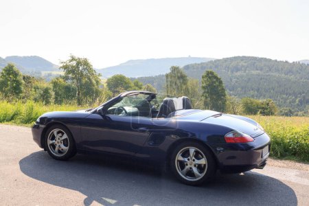 Foto de Gersfeld, Germany - July 23, 2021: Blue roadster Porsche Boxster 986 with Wasserkuppe panorama in Rhoen Mountains. The car is a mid-engine two-seater sports car manufactured by Porsche. - Imagen libre de derechos