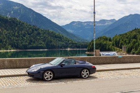 Photo for Lenggries, Germany - July 29, 2023: Blue Porsche 911 convertible parking on Sylvenstein Dam with lake Sylvenstein Reservoir and mountain alps panorama in Bavaria. - Royalty Free Image