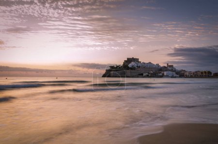 Beautiful landscape of the old town of Peniscola with the castle on the top and the sea waves on the beach in the foreground at sunrise, Peniscola, Castellon, Spain