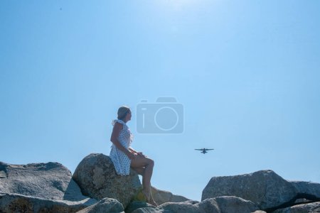 Photo for One girl in a white dress sits on gray rocks and looks at an airplane. High quality photo - Royalty Free Image