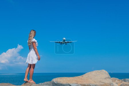 Photo for Meeting of Elements: White Dress, Blue Sea, Stones, and the Flying Plane. High quality photo - Royalty Free Image