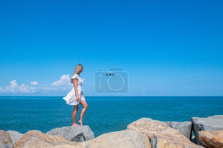 Photo for Beyond the Shore: Girl in White Dress, Stones, Sea, and an Airborne Plane. High quality photo - Royalty Free Image