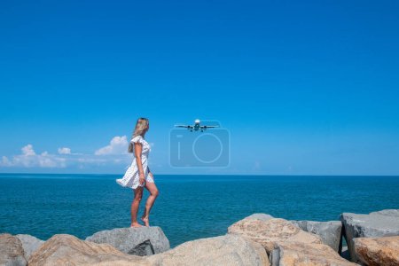 Photo for Coastal Magic: Girl in White Dress on Stones, Meeting a Plane by the Blue Sea. High quality photo - Royalty Free Image