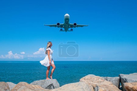 Aerial Serenity: Mädchen in weißem Kleid auf Steinen, Flugzeug, das über dem blauen Meer schwebt. Hochwertiges Foto