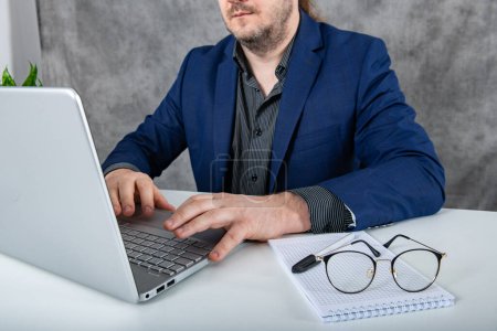 Serious businessman wearing a blue suit sitting at a tidy desk while typing on a keyboard and taking notes in a notepad, depicting professionalism and focus in the workplace.