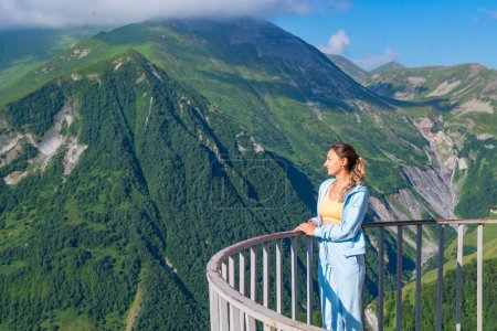 Eine junge Frau im blauen Anzug steht auf einer Aussichtsplattform, im Hintergrund eine majestätische Bergkette. Die Szene strahlt ein Gefühl von Schönheit und Ruhe aus.