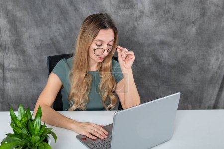 A charming young girl with glasses and a gray dress is gleefully interacting with a laptop while seated next to a verdant green flower, exuding a sense of playful curiosity and joy in learning.