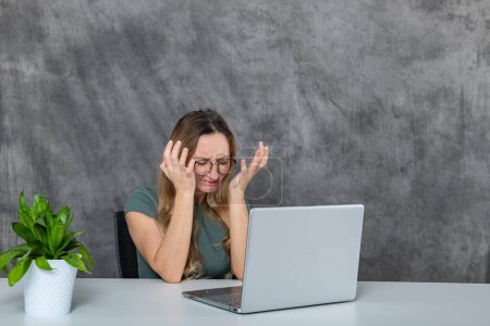 Chica joven con gafas en un vestido gris se ve juguetonamente haciendo caras delante de un ordenador portátil. Una maceta verde vibrante da vida al fondo, mejorando el atractivo general de la imagen.
