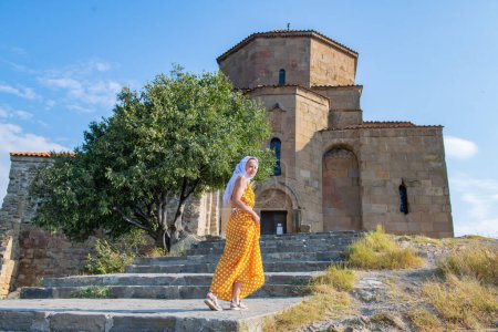 Eine modische Frau in einem leuchtend orangefarbenen Kleid und einem fließenden weißen Schal posiert an einem strahlend sonnigen Tag vor einer malerischen, historischen alten Kirche..