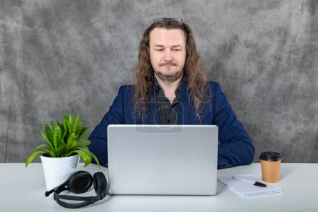 Un homme aux cheveux longs à la mode et confiant dans un costume bleu sophistiqué posant avec un ordinateur portable élégant et des écouteurs à la mode, mettant en valeur la technologie moderne et l'élégance professionnelle.
