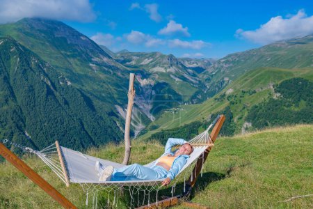 Eine junge Frau im blauen Anzug entspannt in einer Hängematte und genießt die atemberaubende Aussicht auf die Berge im Hintergrund. Sie wirkt ruhig und zufrieden inmitten der Natur.