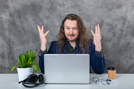 A stylish and confident young man with long hair dressed in a blue suit striking poses by a laptop and various accessories in a modern office setting, showcasing professionalism and confidence.
