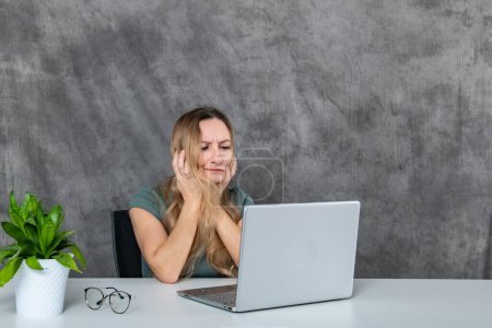 A young woman with lovely blond hair and an elegant gray dress creates various poses in front of a laptop, with a vibrant green flower adding a touch of nature to the scene.