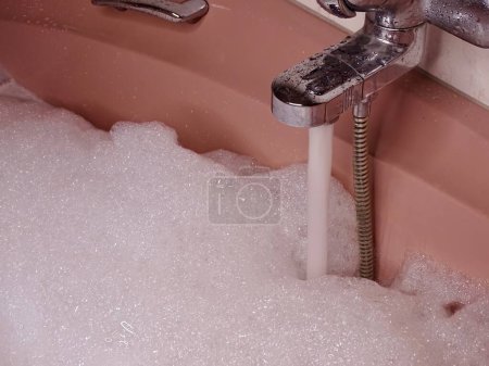 Bañera rosa llena hasta el borde: flujos de grifo metálicos con agua y espuma abundante